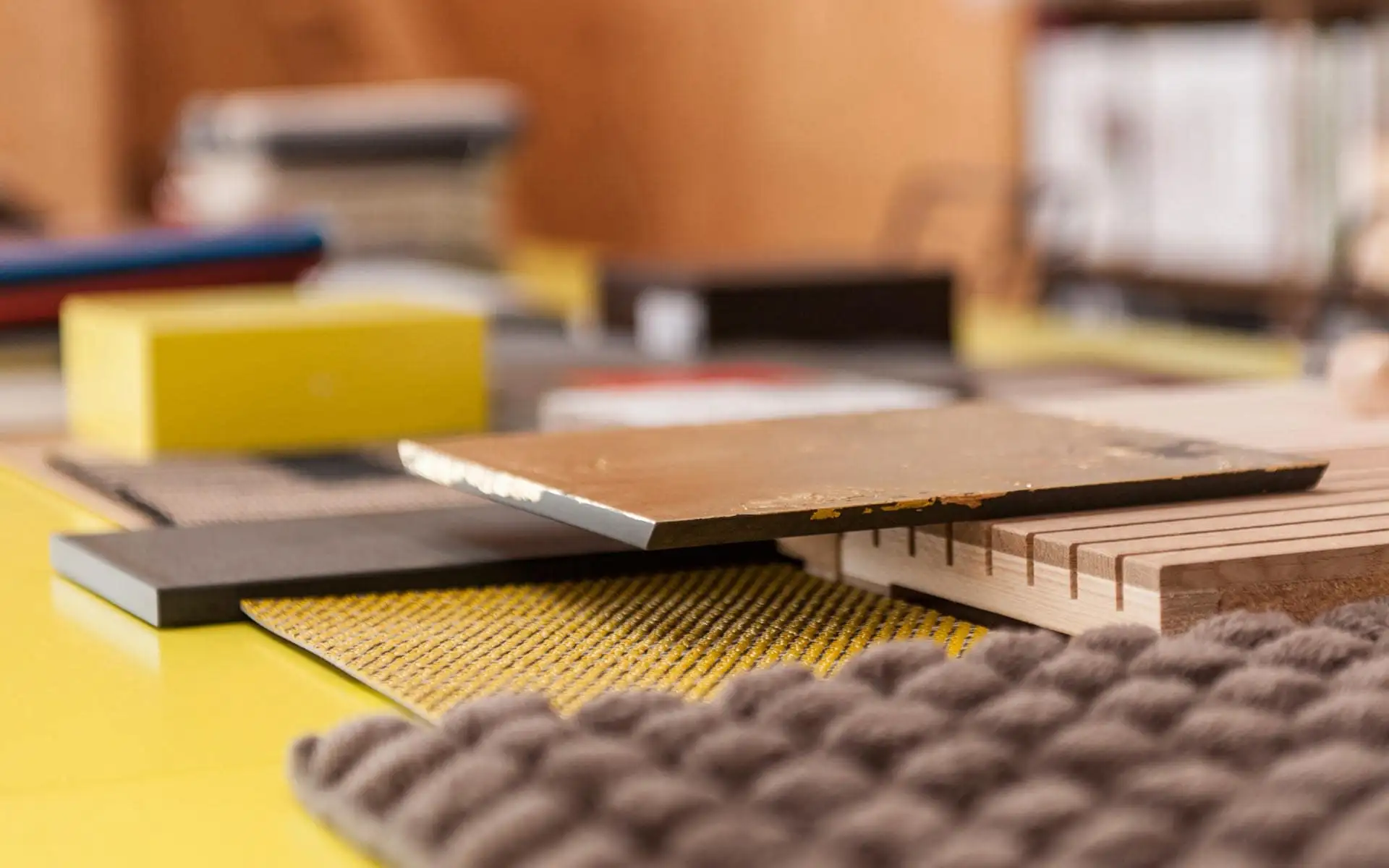 Arbeitsfläche mit Materialmustern aus Holz, Metall und Textil; im Vordergrund eine gefräste Holzprobe mit eingesetzten Nägeln.