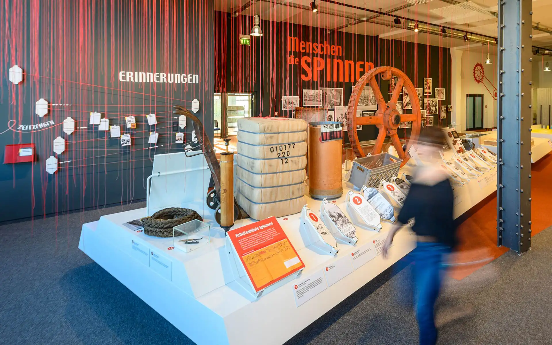 Ausstellungsbereich mit roten Fäden, historischen Objekten und dem Schriftzug Menschen die spinnen