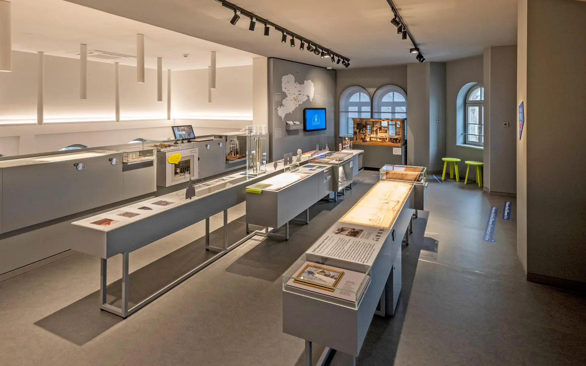 Ausstellungsraum mit langen Vitrinentischen, Sachsen-Wandgrafik und Medienstation; ruhige, helle Beleuchtung.