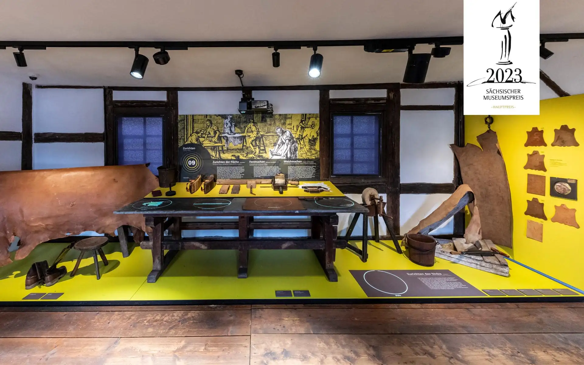 Gerberwerkstatt, Werkbank mit Werkzeugen, gelbe Tafeln, historische Zeichnung im Hintergrund