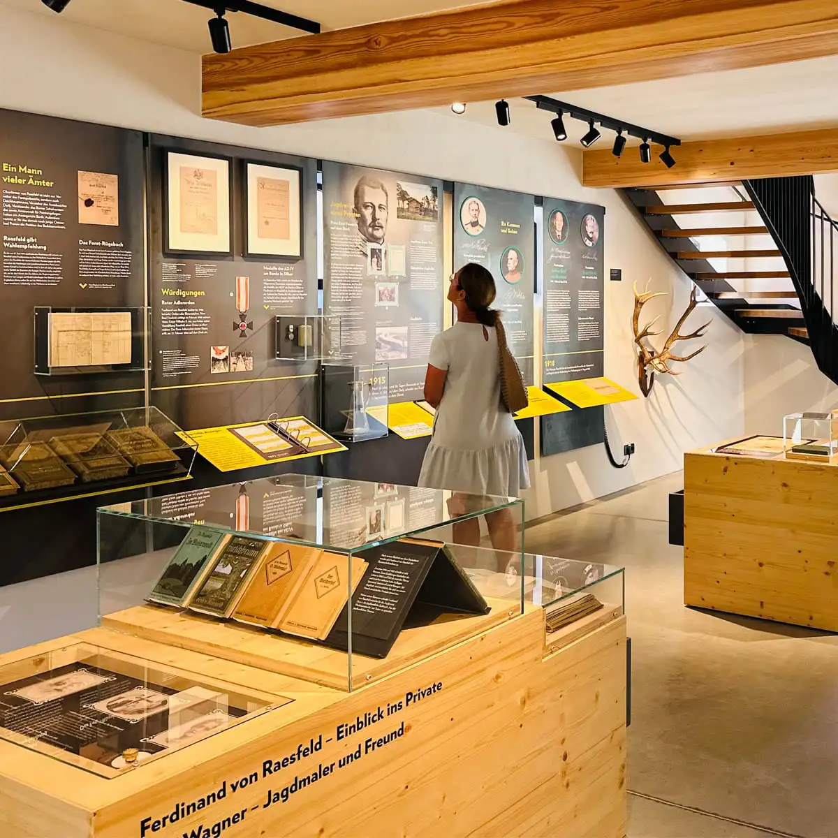 Besucherin vor Wandtafeln mit Fotos und Texten; davor Vitrinen mit Büchern und Dokumenten, Treppe mit Geweih an der Wand.