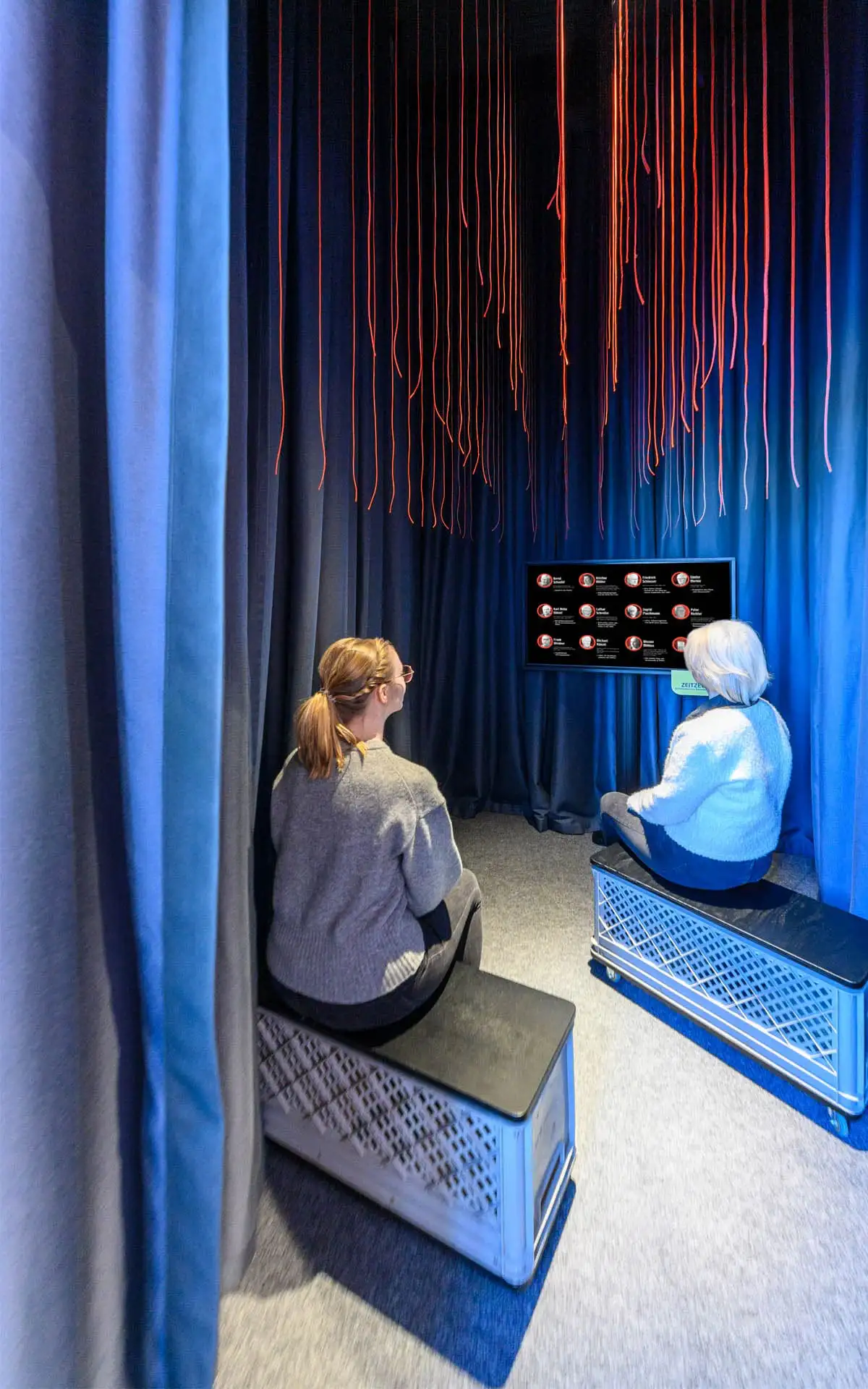 Zwei Besucherinnen sitzen in einer dunklen Hörstation vor einem Bildschirm unter herabhängenden roten Fäden