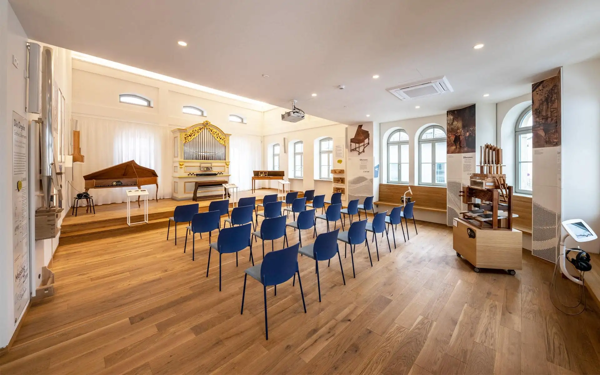 Musiksaal mit Orgel und Cembalo, Reihen blauer Stühle, Parkettboden und Rundbogenfenster.