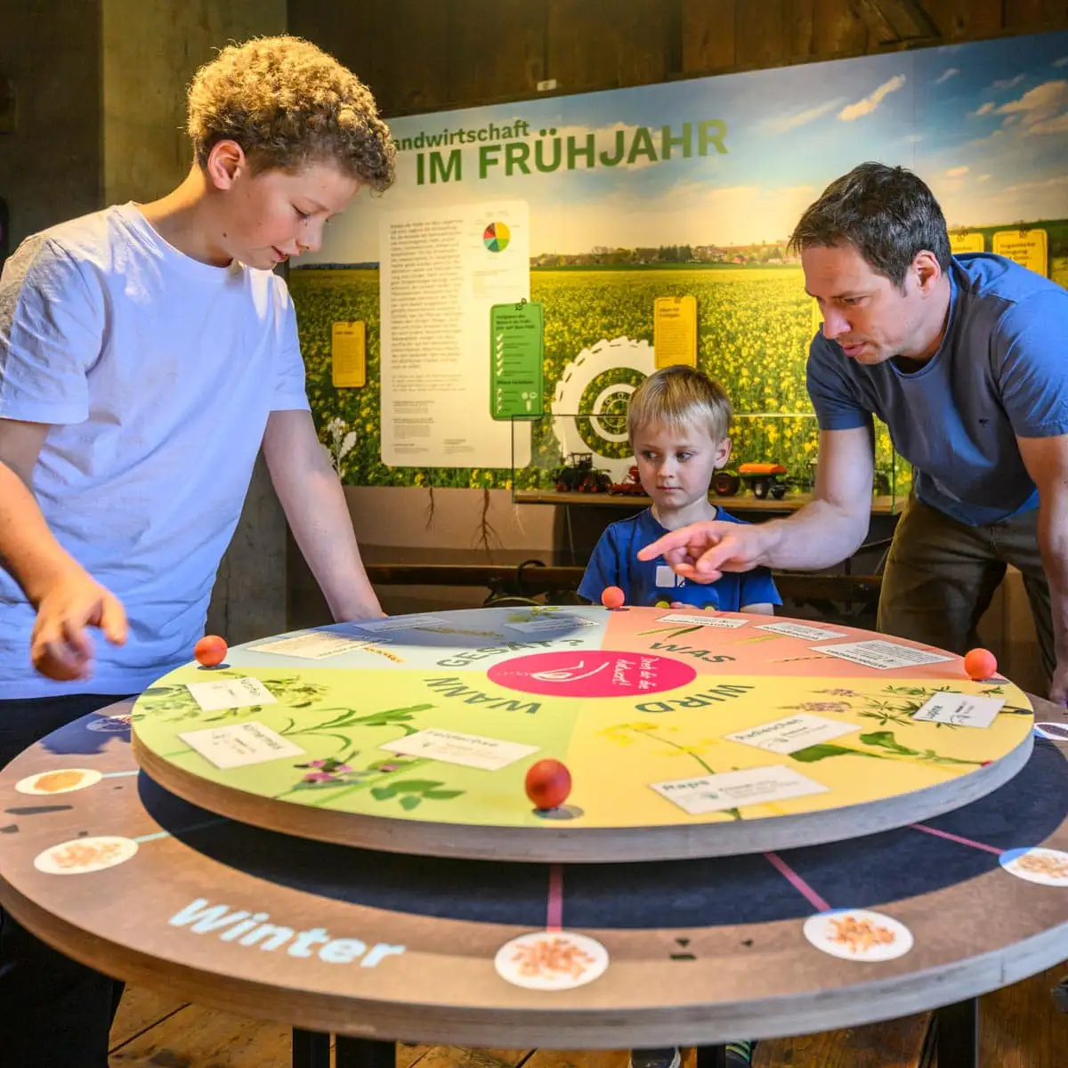 Interaktive runde Lernstation, zwei Kinder und ein Erwachsener bedienen eine drehbare Scheibe