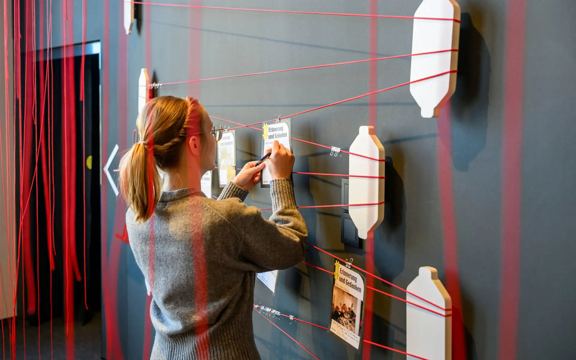 Besucherin befestigt eine Karte an einer Erinnerungswand mit gespannten roten Fäden