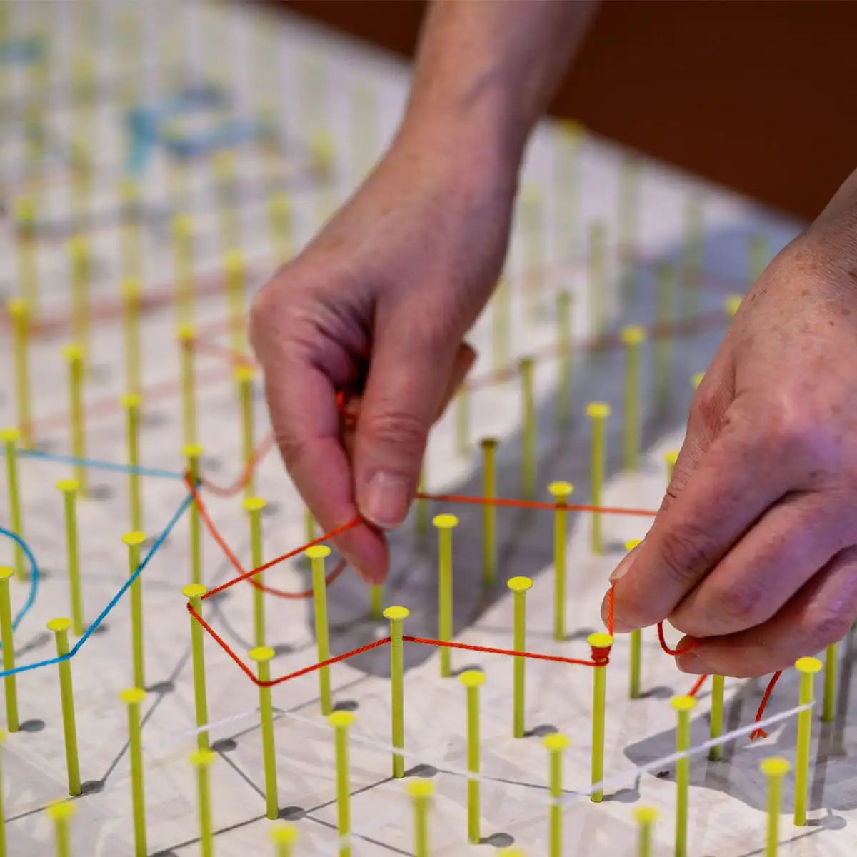 Hände spannen orangefarbenen Faden über eine interaktive Station mit Steckpunkten