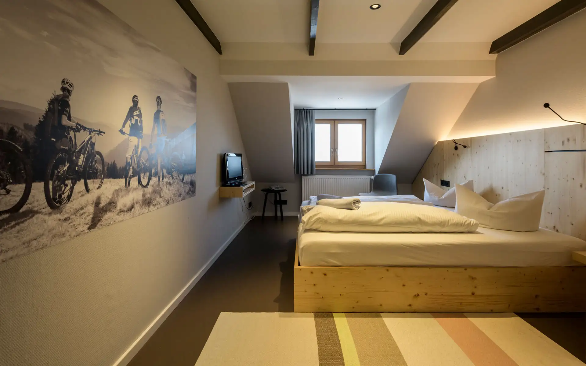 Blick entlang des Hotelzimmers mit Holzbett, großem Wandbild mit Mountainbikern und Fenster an der Stirnseite.
