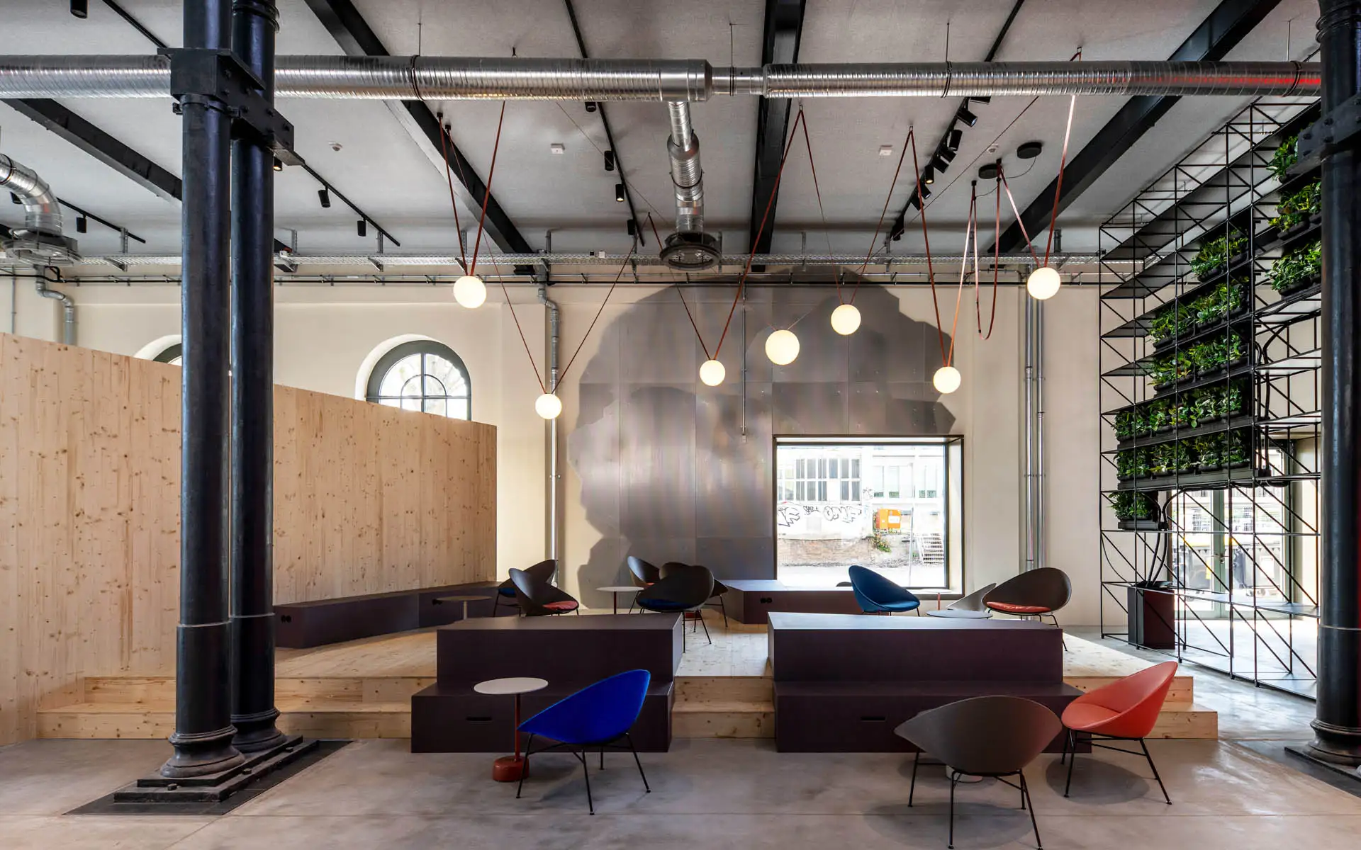 Sitzbereich mit farbigen Sesseln, runden Leuchten und großem Wandrelief in einer offenen Industriehalle.
