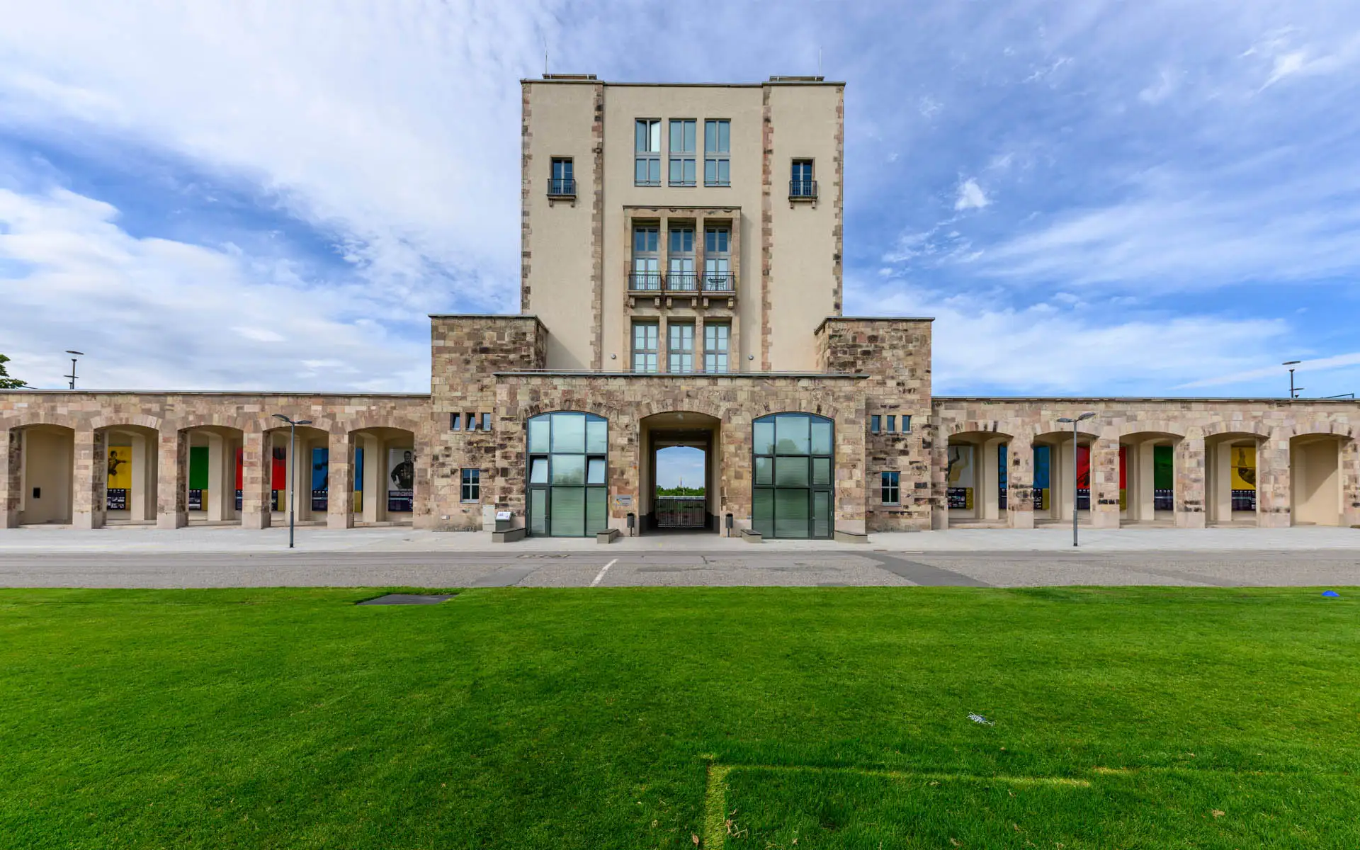 Sportforum Chemnitz mit Turm, Arkaden und grüner Rasenfläche unter blauem Himmel.