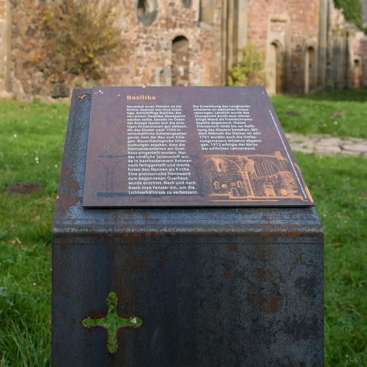 Tafel mit Text zur Basilika auf einem Sockel aus Cortenstahl vor der Kirchenruine