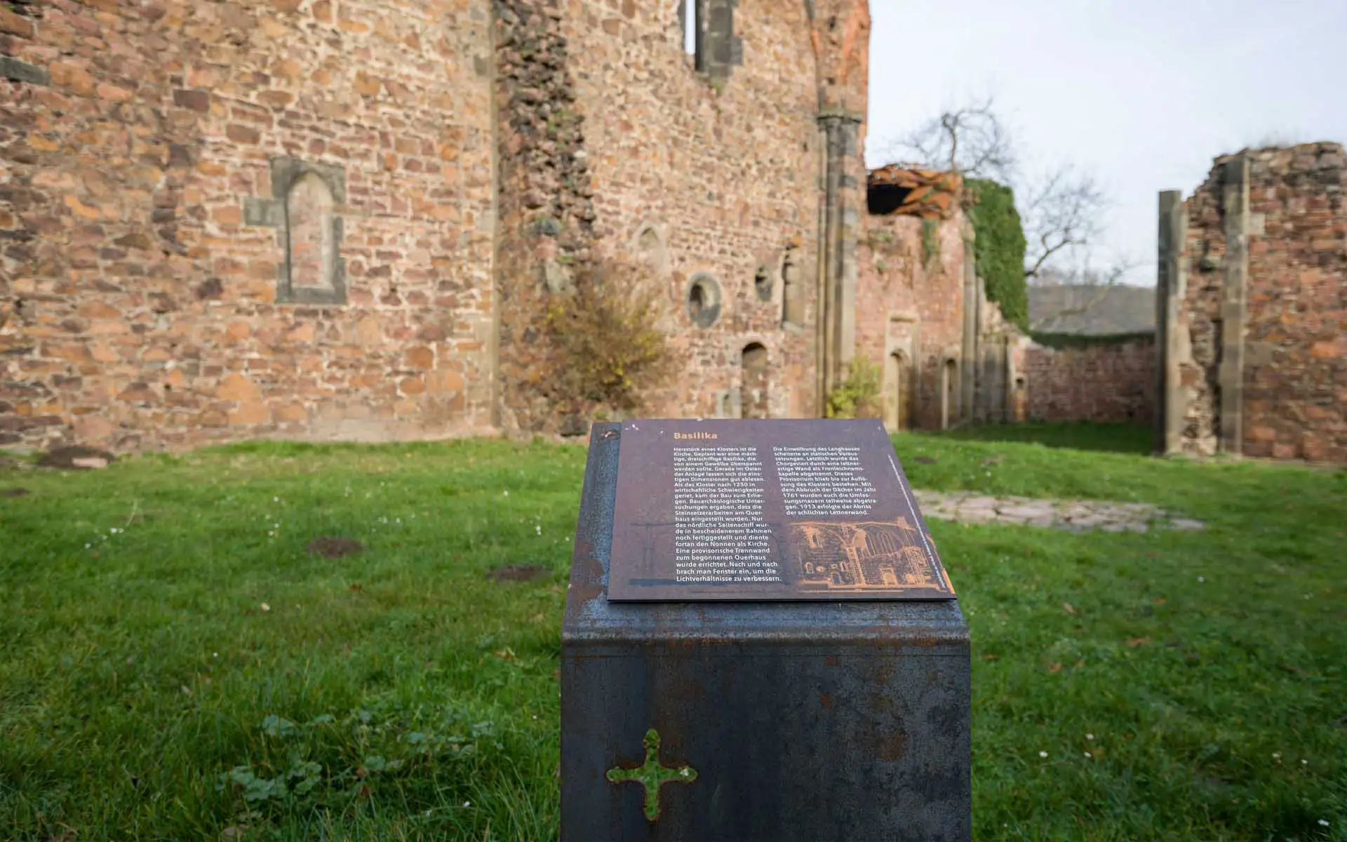 Infotafel vor der historischen Heilig-Kreuz-Ruine, mit Text zur Basilika und rostfarbener Metallstele.