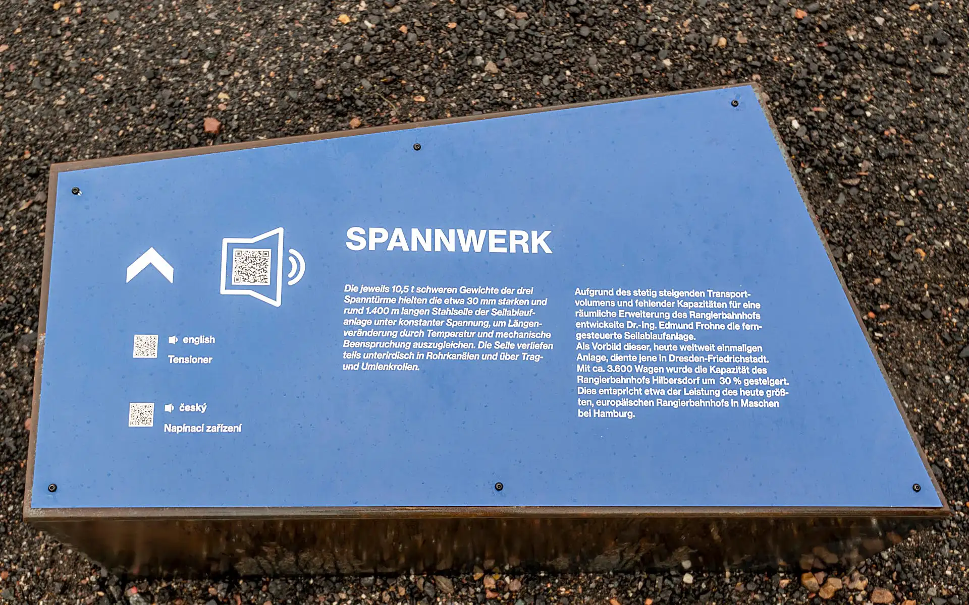 Blaue Infotafel 'Spannwerk' mit erklärendem Text und QR-Codes, auf Kiesboden.