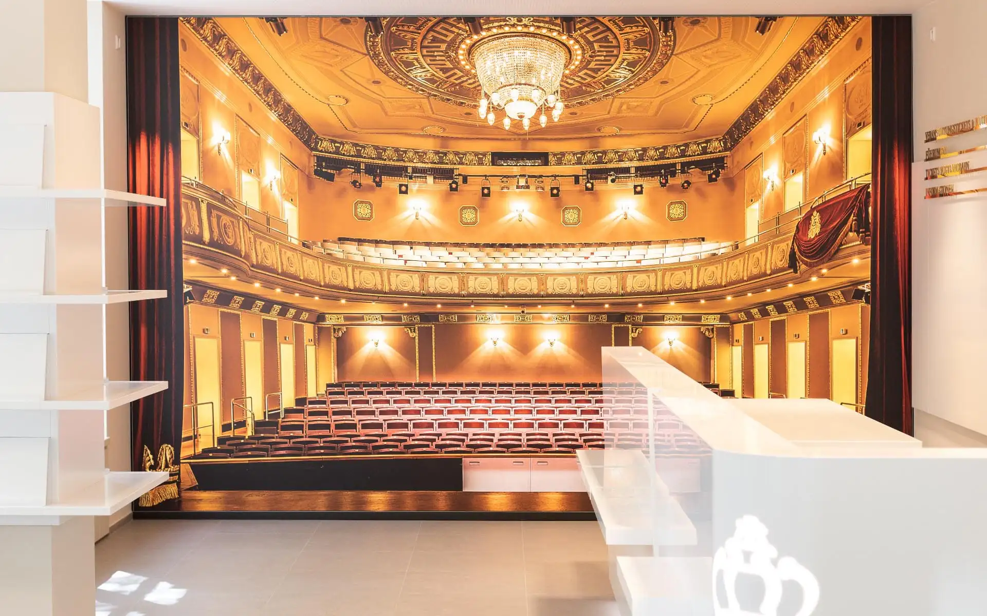 Tourist-Information mit weißem Tresen vor großformatigem Wandbild eines Theatersaals.