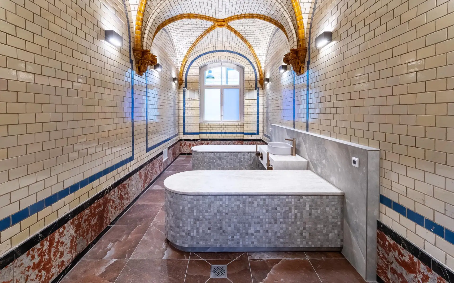 Historischer Gewölberaum mit cremefarbenen Fliesen, blauen Zierlinien und zwei Mosaik-Wannen