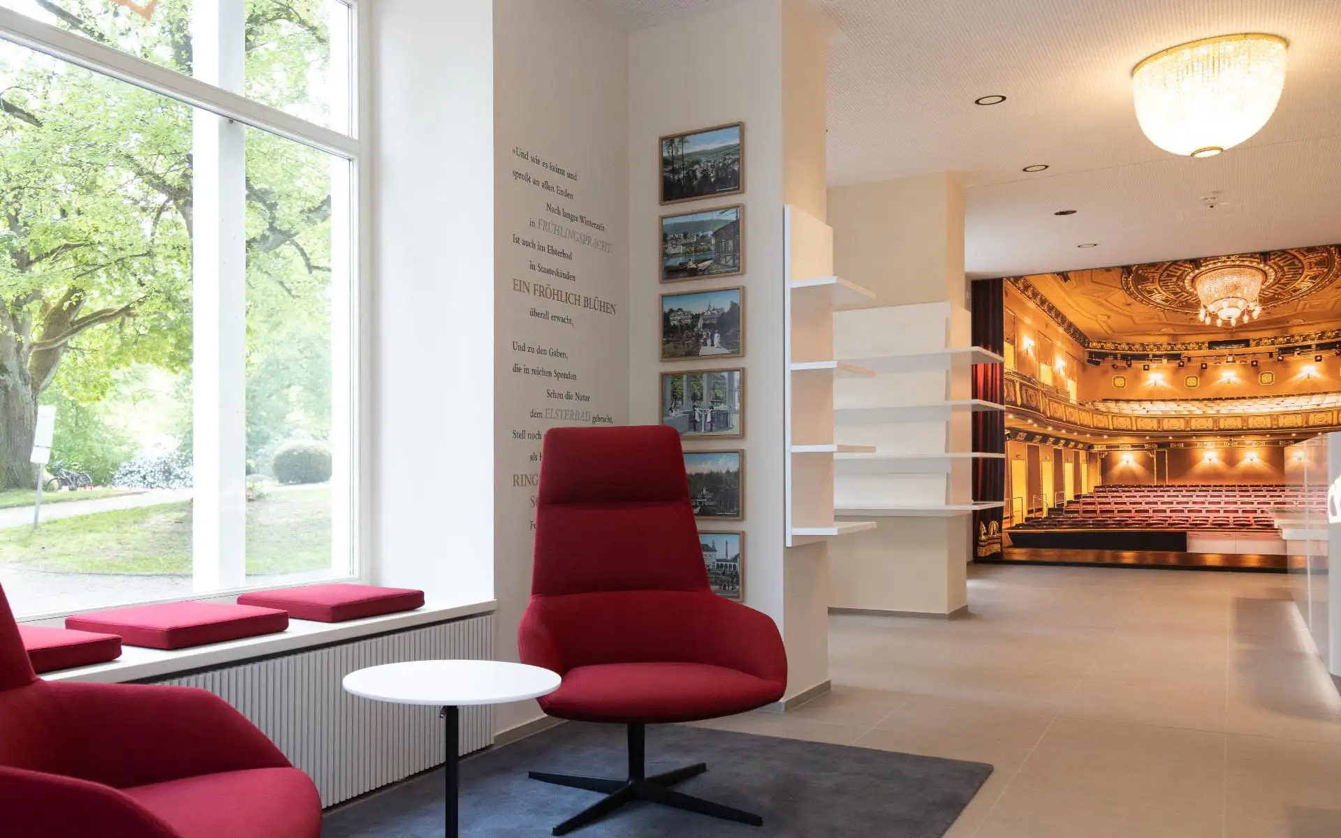 Sitzecke mit roten Sesseln am Fenster; im Hintergrund Empfangsbereich mit großem Theater-Wandbild.