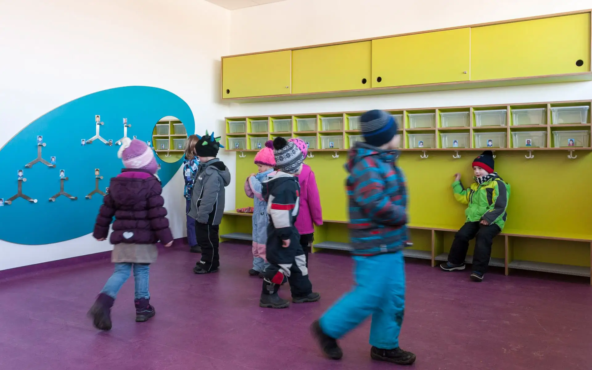 Kinder in Winterkleidung im Garderobenbereich vor Fächern und Hakenleisten