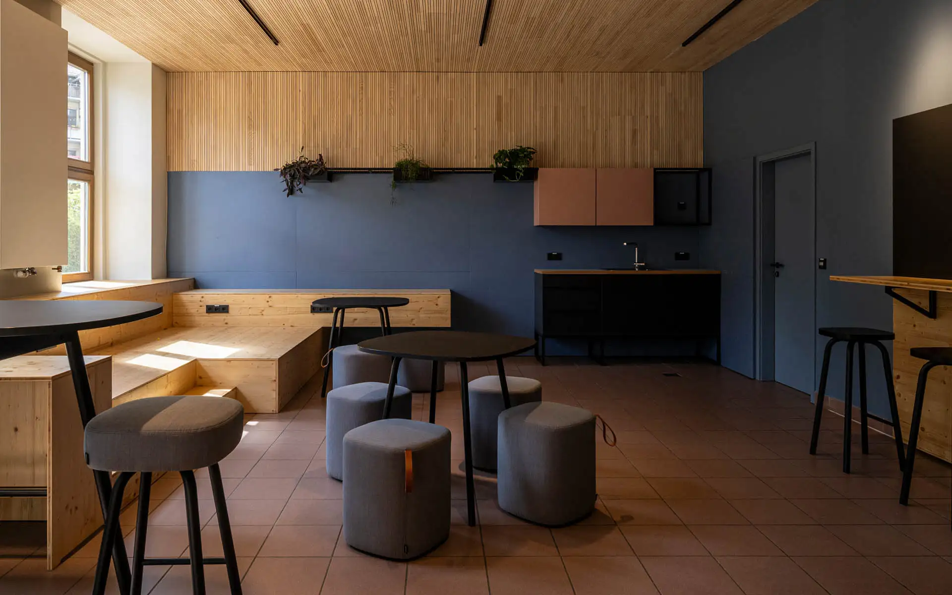 Aufenthaltsraum mit Holzpodest, runden Tischen und Hockern; blaue Wand mit Küchenzeile.