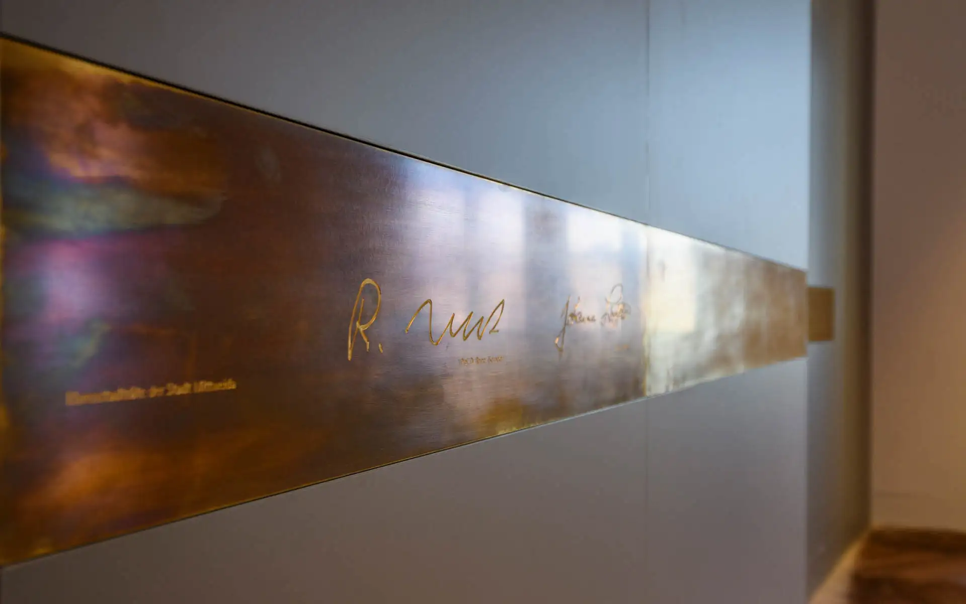 Detail einer eingelassenen Metallleiste mit gravierten Unterschriften an der Saalwand