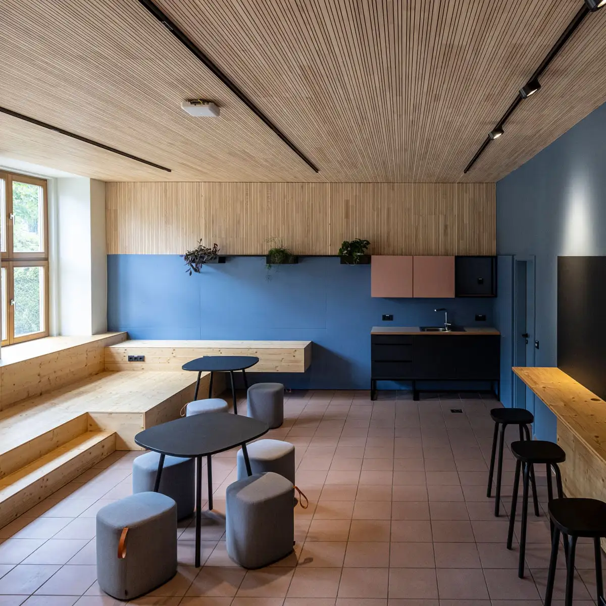 Raumansicht mit Holzlamellendecke, Sitzstufen und Küchenzeile vor blauer Wand.