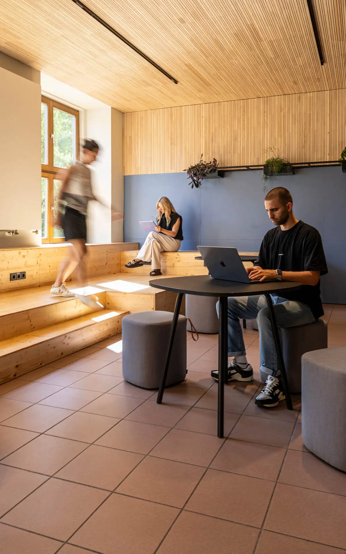 Person arbeitet am Laptop am Tisch; im Hintergrund sitzt eine weitere Person auf den Holzstufen, eine läuft vorbei.
