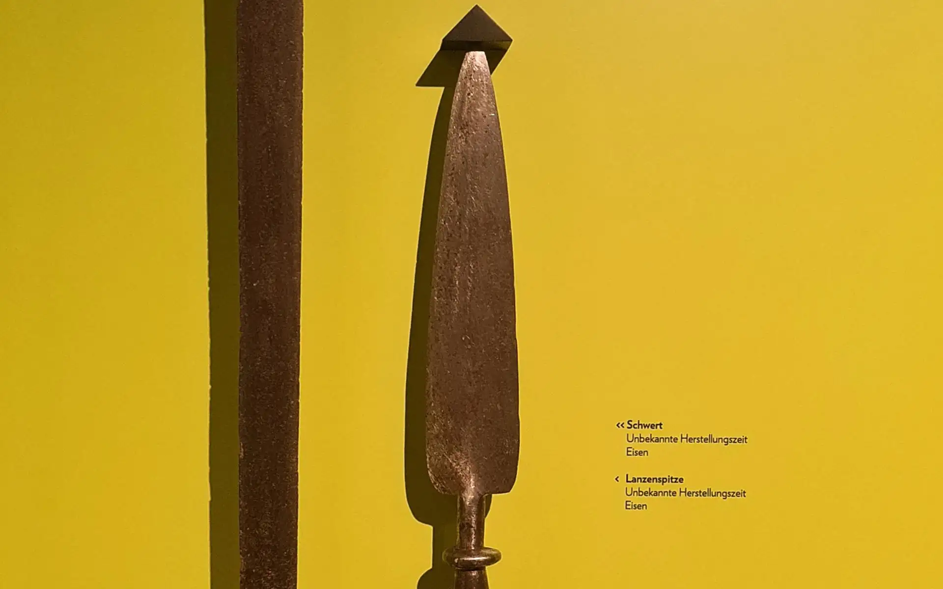 Historische Waffenexponate wie Schwert und Lanzenspitze vor gelbem Hintergrund mit erklärender Beschriftung.