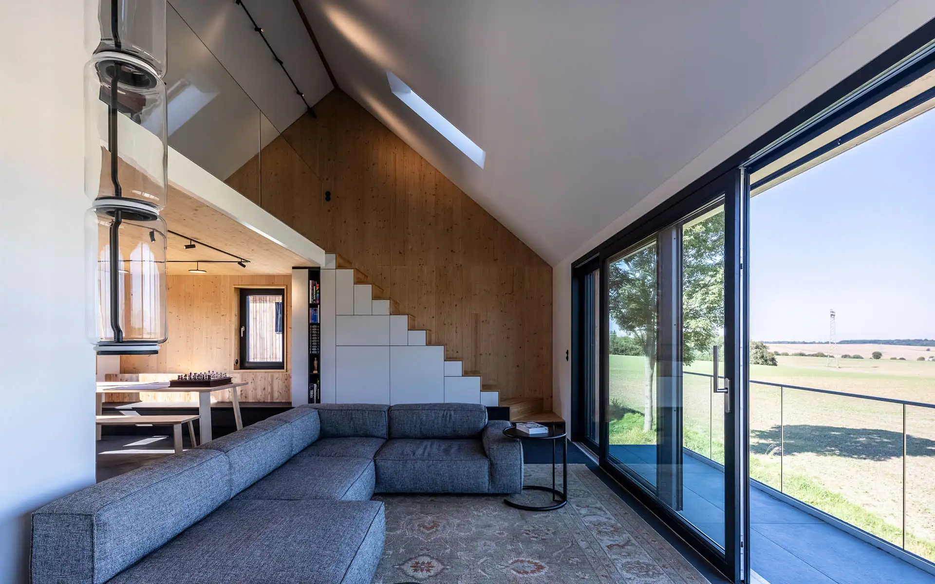 Offener Wohnraum mit Sofa, Galerieebene, Holzverkleidungen und großflächiger Verglasung mit Blick in die Landschaft.