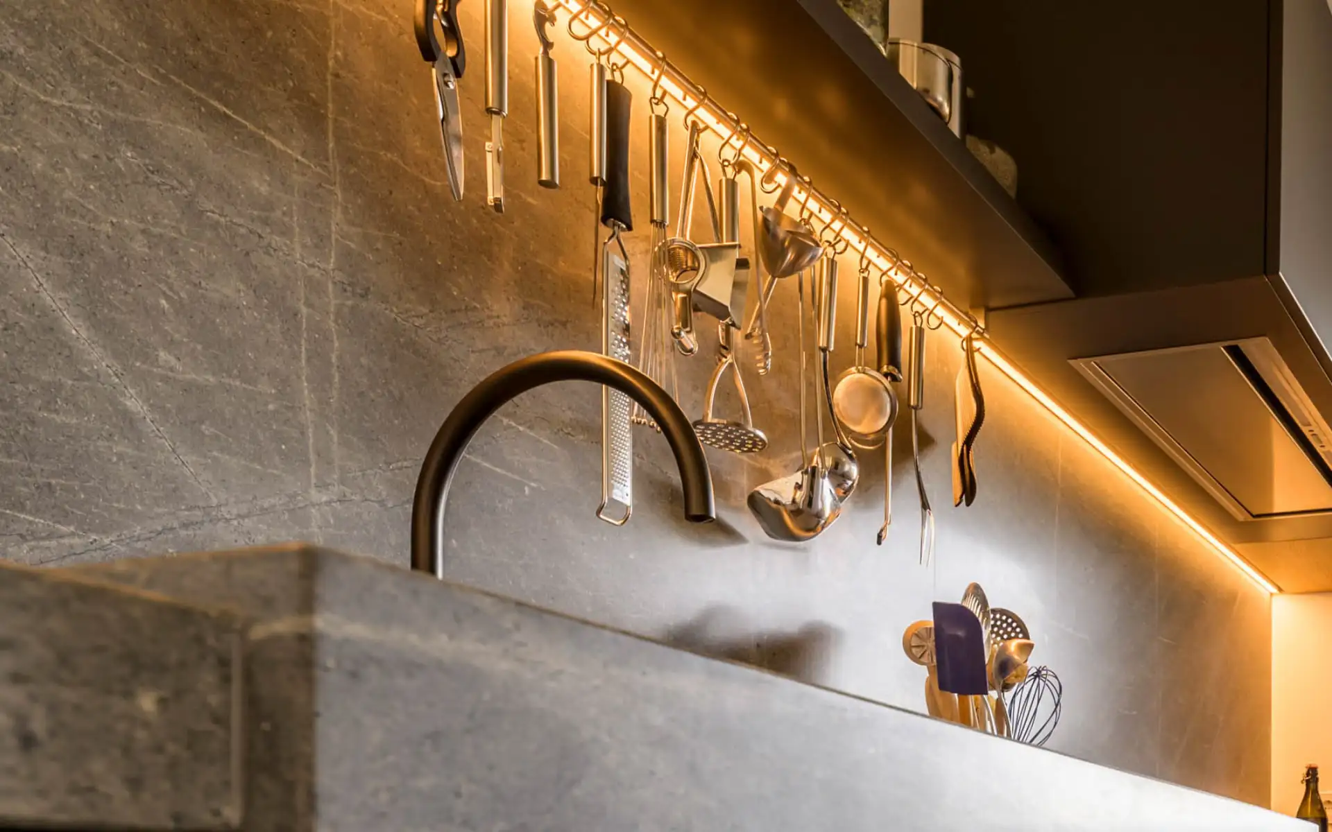 Küchenrückwand aus Naturstein mit LED-Lichtleiste, Hängeschiene und mattschwarzer Armatur.