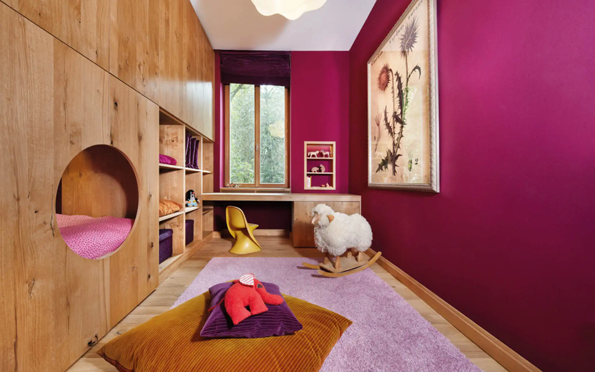 Kinderzimmer mit magenta Wänden, Einbaumöbeln aus Eiche und runder Schlafnische. Schreibtisch am Fenster, gelber Stuhl und Schaukelschaf.