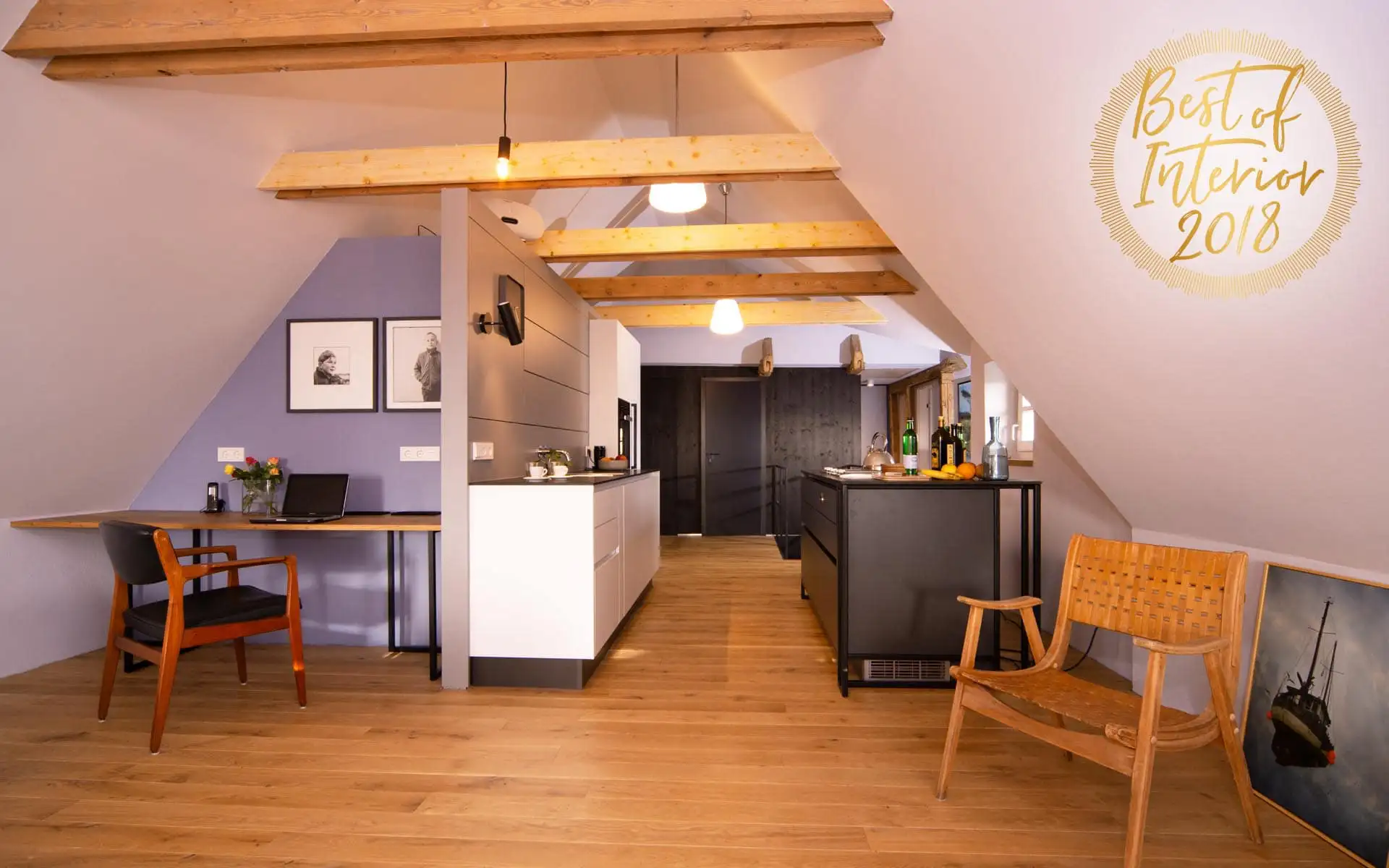 Dachgeschoss mit sichtbaren Holzbalken: links Arbeitsplatz, mittig weiße Küchenzeile, rechts Kücheninsel und Holzstuhl.