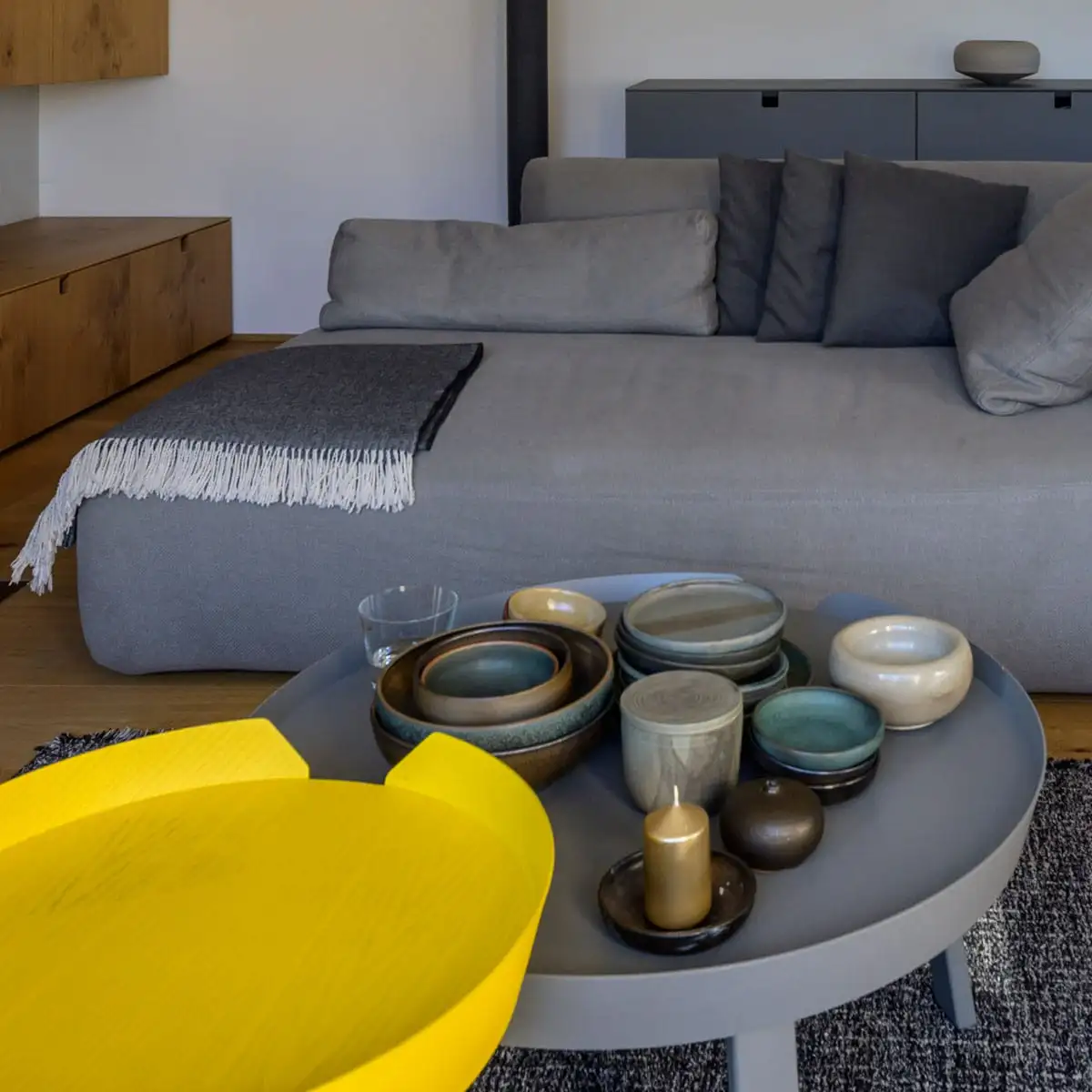 Detail: Couchtisch mit Keramikschalen und gelbem Tablett vor grauem Sofa.