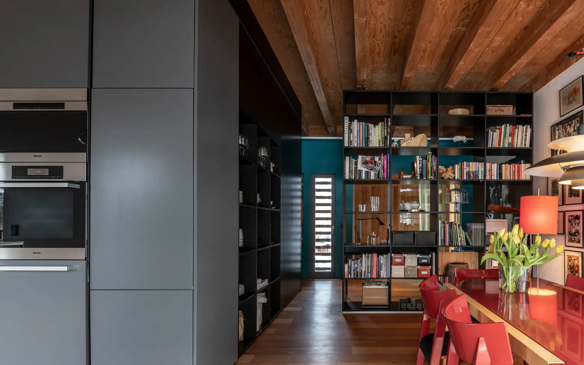 Essbereich mit rotem Tisch und Stühlen, große schwarze Regalwand mit Büchern und Objekten; Holzbalkendecke.