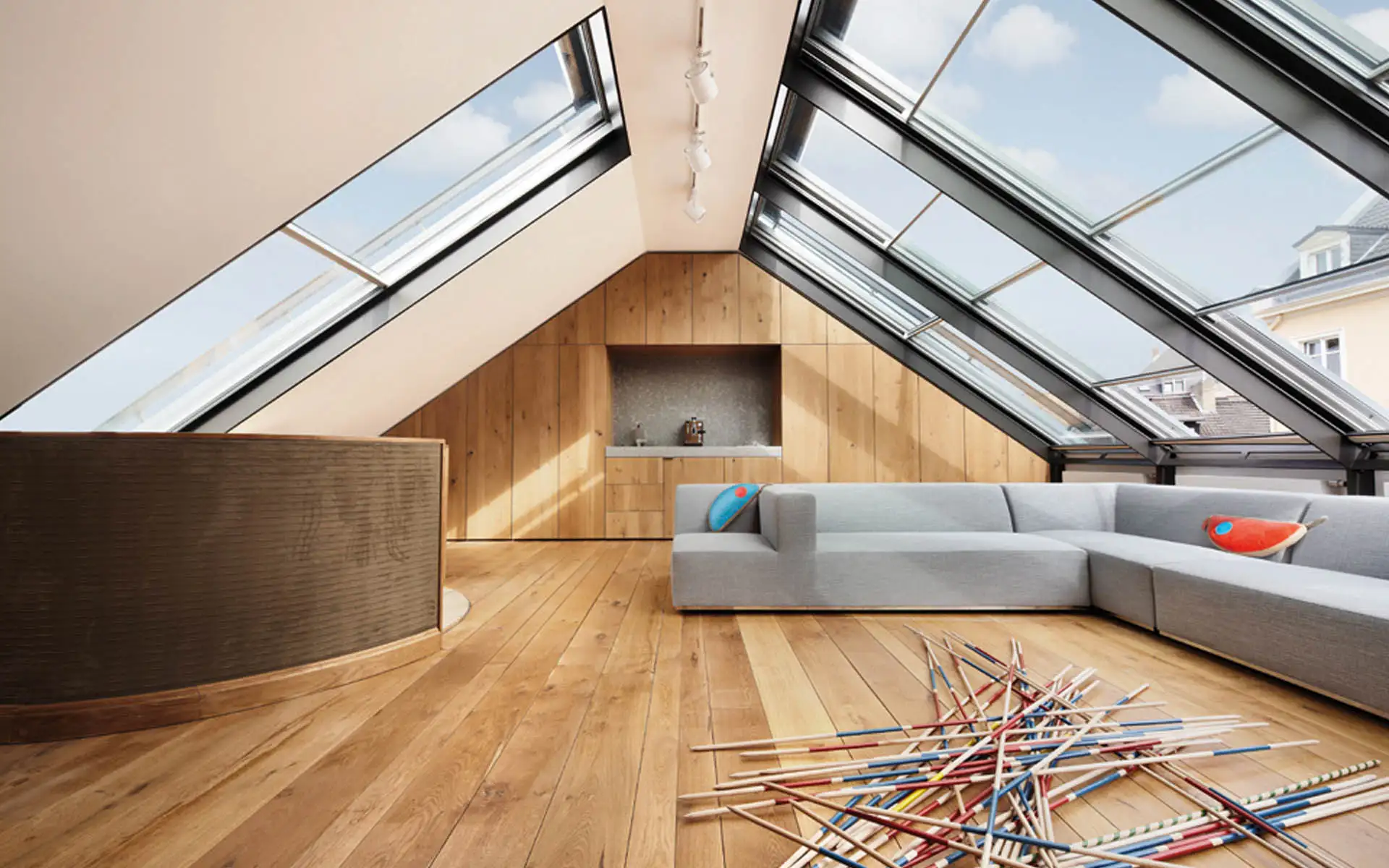 Dachstudio mit großflächigen Dachfenstern, Eichenverkleidung und grauem Sofamodul; Mikado-Stäbe auf dem Parkett.