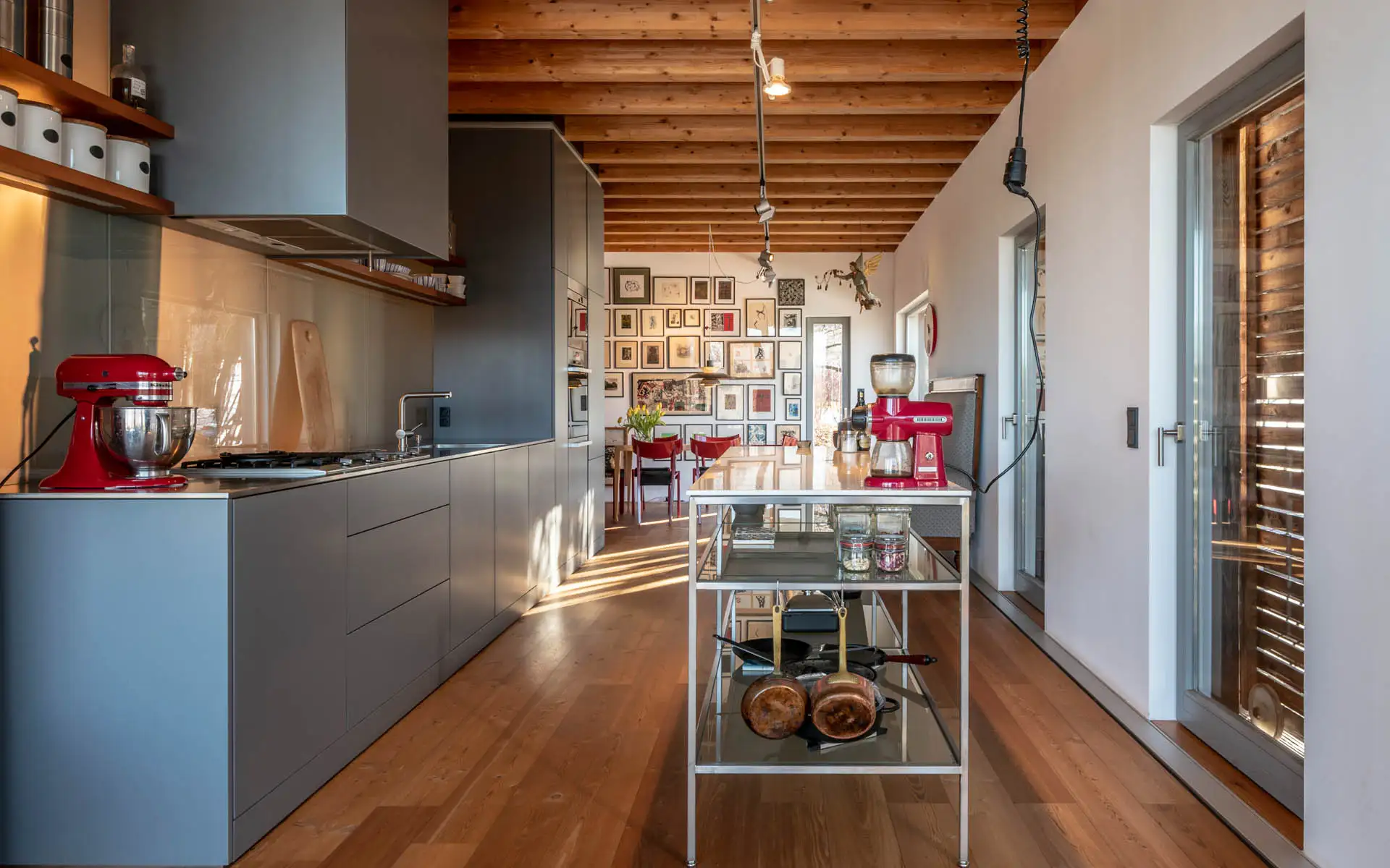 Lange Küchenzeile in Grau, mobile Insel aus Edelstahl, rote Geräte; Blick zur gerahmten Bilderwand im Hintergrund.