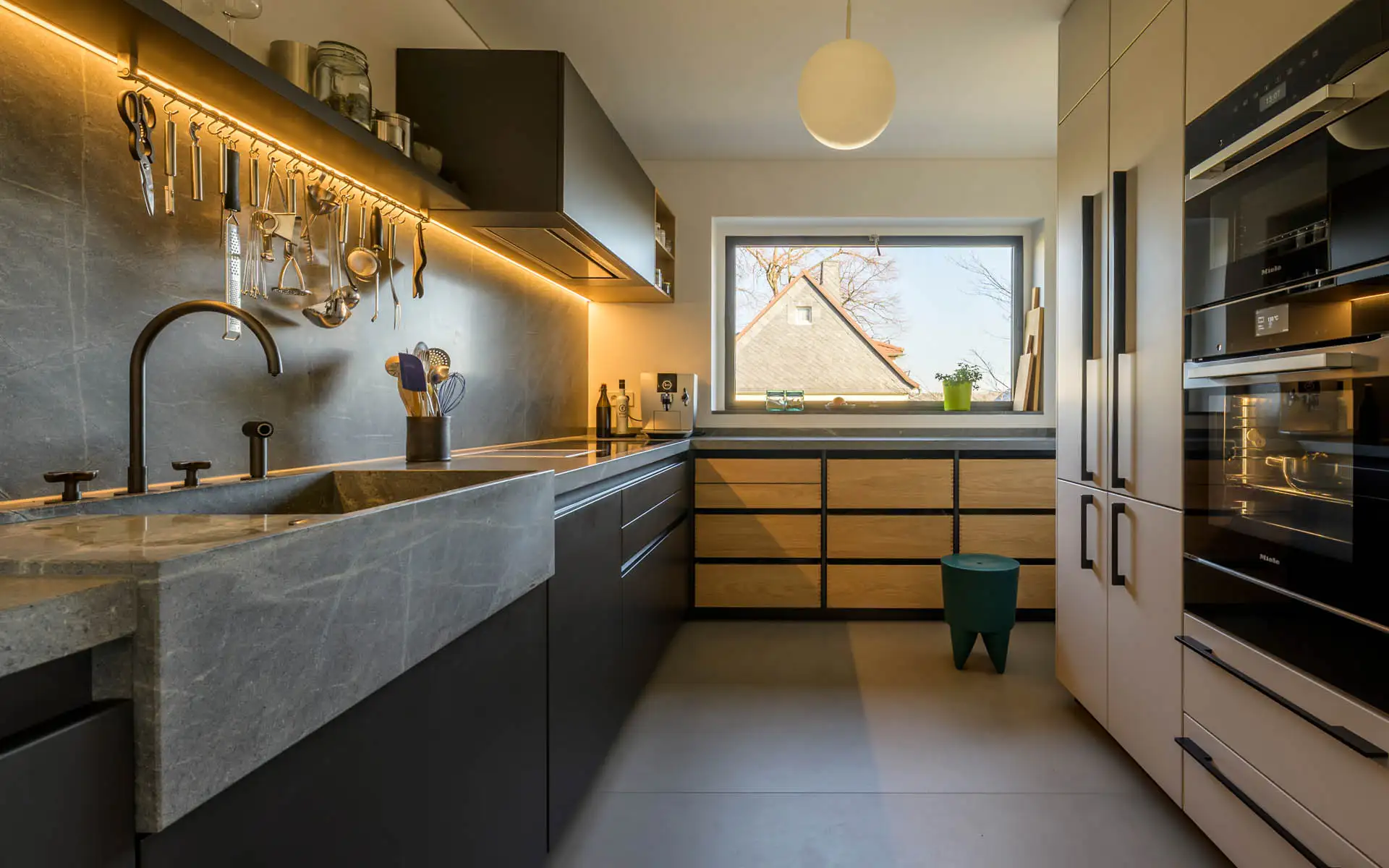 U-förmige Küche mit Naturstein, Gerätehochschrank und Fenster; warm akzentuierte Beleuchtung.