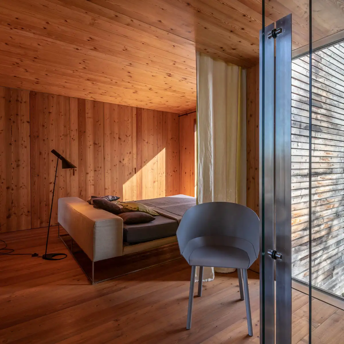 Schlafzimmer mit Wänden und Decke aus Holz, Tageslicht durch Glasfront; grauer Stuhl vorn, Bett im Hintergrund.