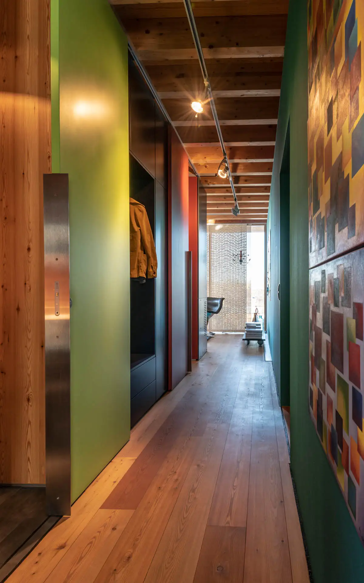 Schmaler Flur mit farbigen Paneelen, Einbauten und Sicht auf einen Arbeitsplatz am Ende.